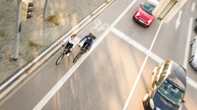 Zwei Fahrradfahrer:innen im Straßenverkehr mit Autos, von schräg vorne/oben fotografiert