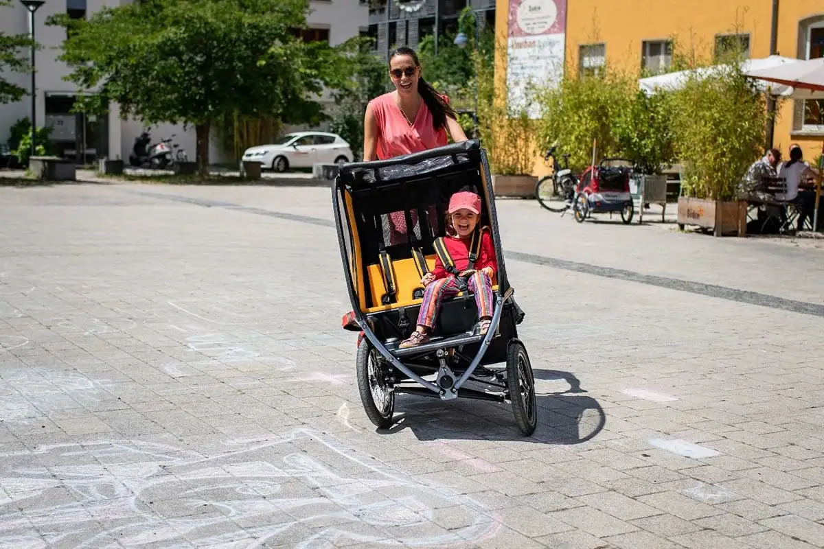 Eine Mutter fährt das Chike e-Bike in der Stadt. Ihr Kind sitzt angeschnallt darin. Beide lachen Eine Mutter fährt das Chike e-Bike in der Stadt. Ihr Kind sitzt angeschnallt darin. Beide lachen